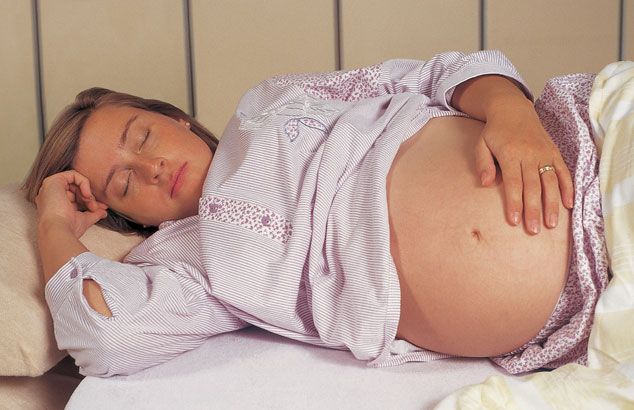 A heavily pregnant woman sleeps on her side in bed with one hand on her belly and the other by her face.