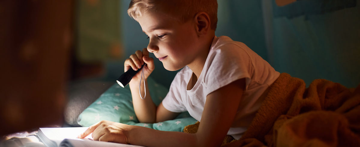 A boy reading a book with a flashlight in his hand while lying in bed with blanket